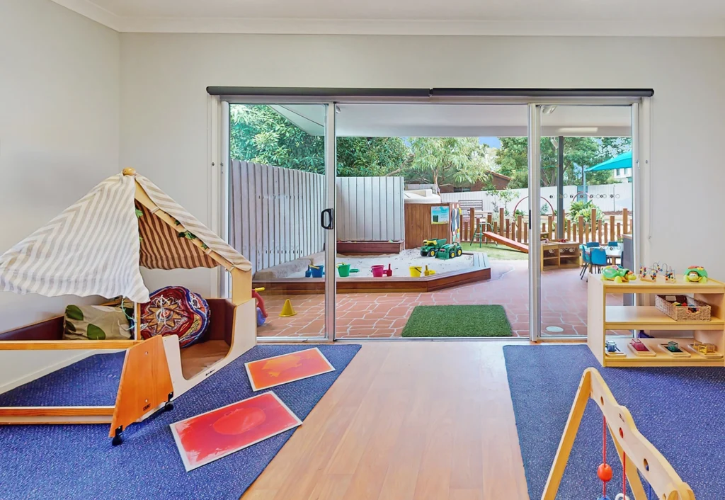 Nursery children's playroom at Busy Bees Underwood Childcare with large sliding glass doors that opens to an outdoor play area with a sandpit.