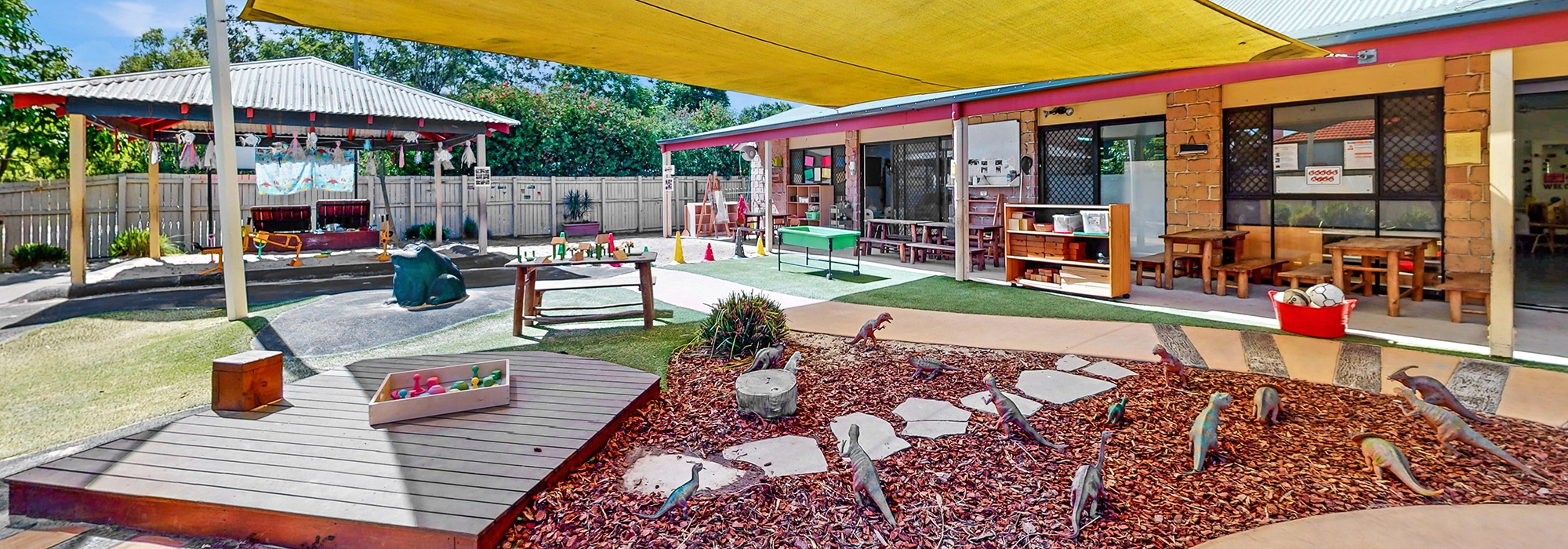 The outdoor area of our early learning center features play equipment, toy dinosaurs, a sandpit, shaded areas, and various activity stations all under a large yellow canopy.