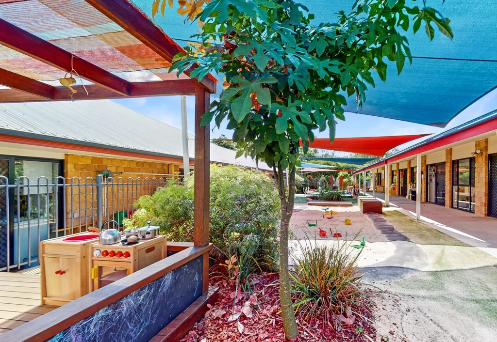 Outdoor playground with a play kitchen, shaded areas, and colorful toys. A fence and building structure are visible in the background, perfect for a fun-filled kindergarten or day care setting.