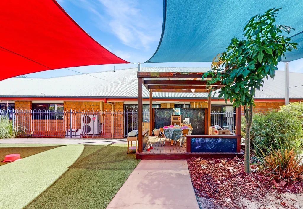 A wooden play structure with a roof stands in an outdoor area featuring artificial turf, perfect for early learning and day care activities. Shaded by colorful fabric canopies, the area is filled with toys and various play items set up under the structure.