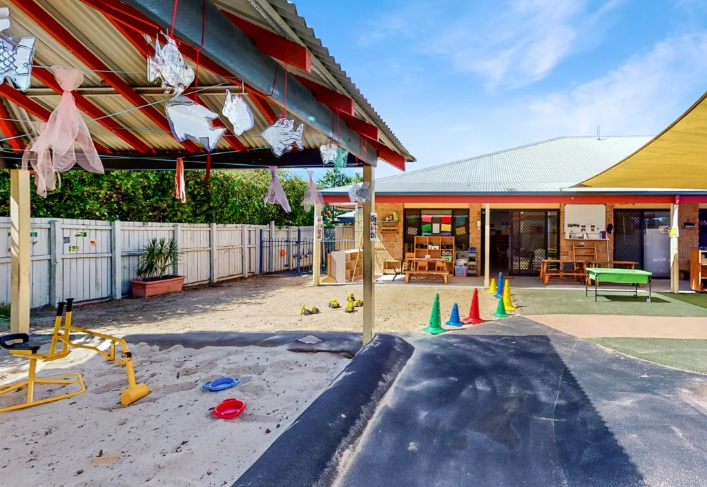 Outdoor play area with a sandpit, colorful cones, and various play equipment designed for preschool fun. An adjacent building with a covered patio is visible, along with chairs and tables. It's a sunny day at this vibrant early learning center.