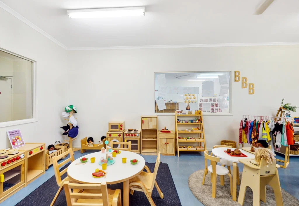 A brightly lit children's playroom in the day care with small tables, chairs, toys, dress-up clothes, and educational materials neatly arranged. The room has white walls, a blue floor, and shelving units for storage.