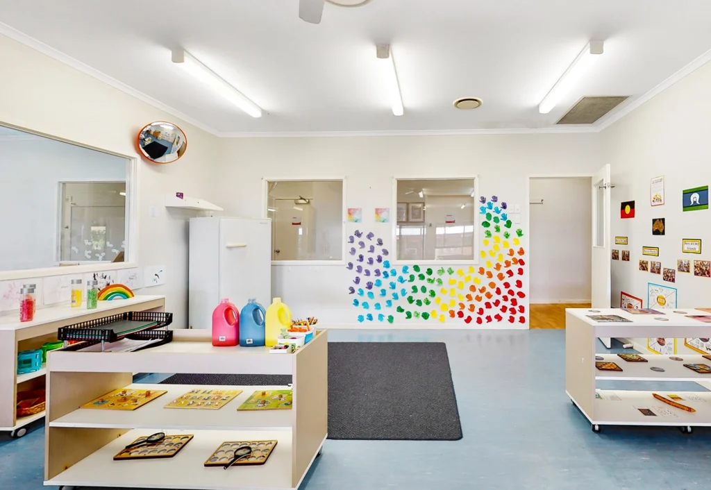 A brightly lit preschool classroom with colorful handprints on the back wall, open white cabinets, and shelves brimming with various educational materials and toys. Two doors and large windows are visible in the background, creating an inviting space for early learning.