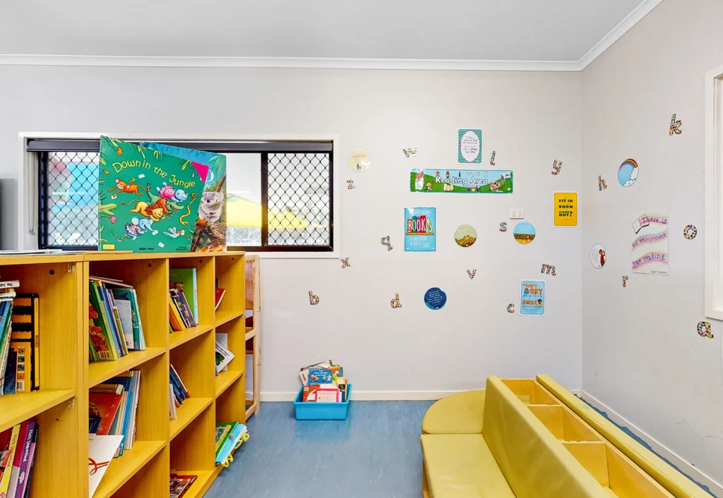 A brightly lit early learning reading corner featuring bookshelves, books, vibrant wall decorations, and cushioned seating.