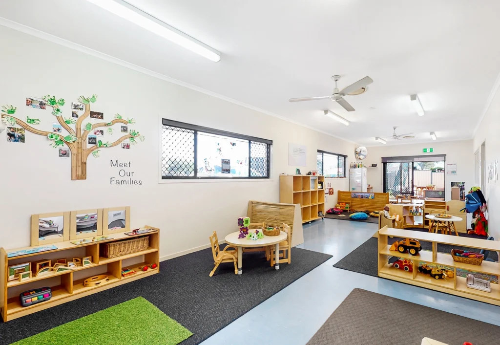 This bright and orderly childcare classroom features various play and learning areas. The space includes small tables, chairs, shelves with toys and books, and a wall display of family photos, creating an inviting environment akin to a warm kindergarten setting.