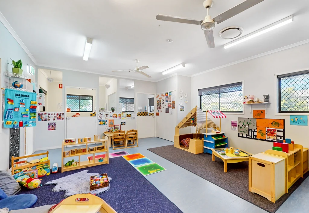 A brightly lit children's playroom, perfect for preschool and kindergarten kids, with toys, books, and various activity stations. The room boasts colorful decorations on the walls and ceiling fans overhead.