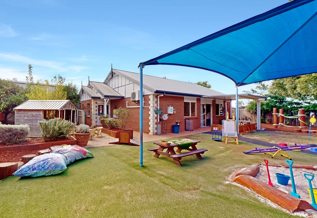 A playground with a shade canopy at the day care features a picnic table, colorful shovels, a small shed, and a brick building in the background, surrounded by greenery.