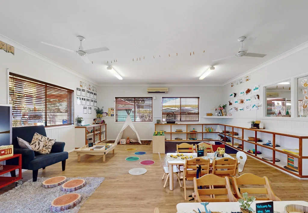 A well-lit, organized children's playroom featuring toys, a table with chairs, a tent, and various shelves with early learning materials, along with colorful decorations on the walls.