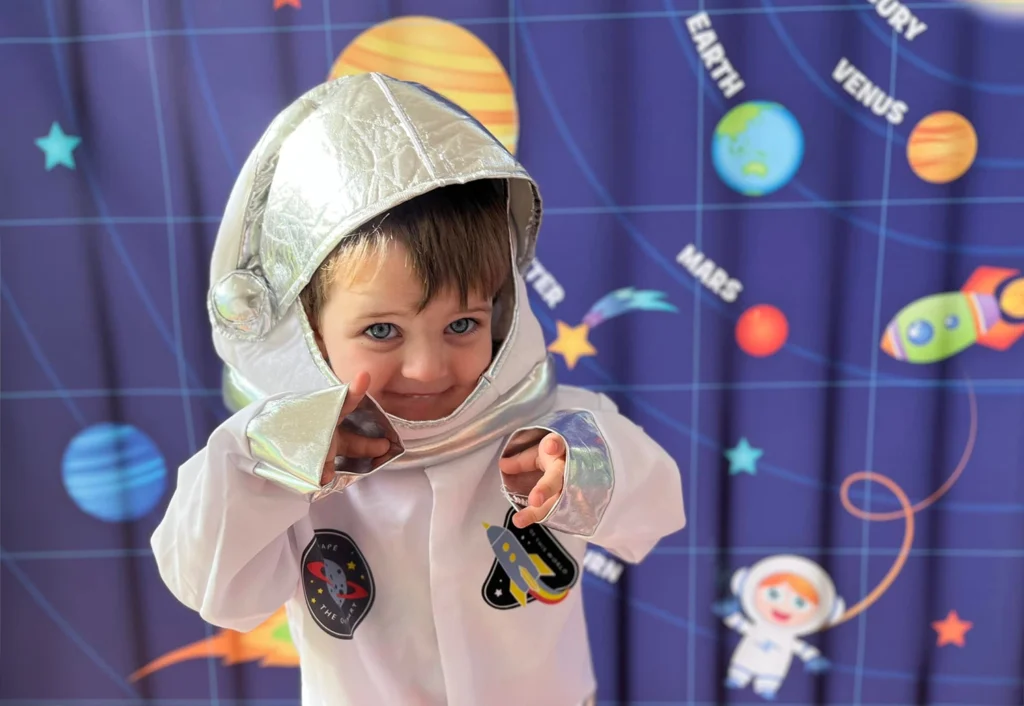 A child dressed as an astronaut stands in front of a space-themed backdrop featuring planets and a rocket ship, capturing the essence of early learning through imaginative play at a high-quality childcare center.