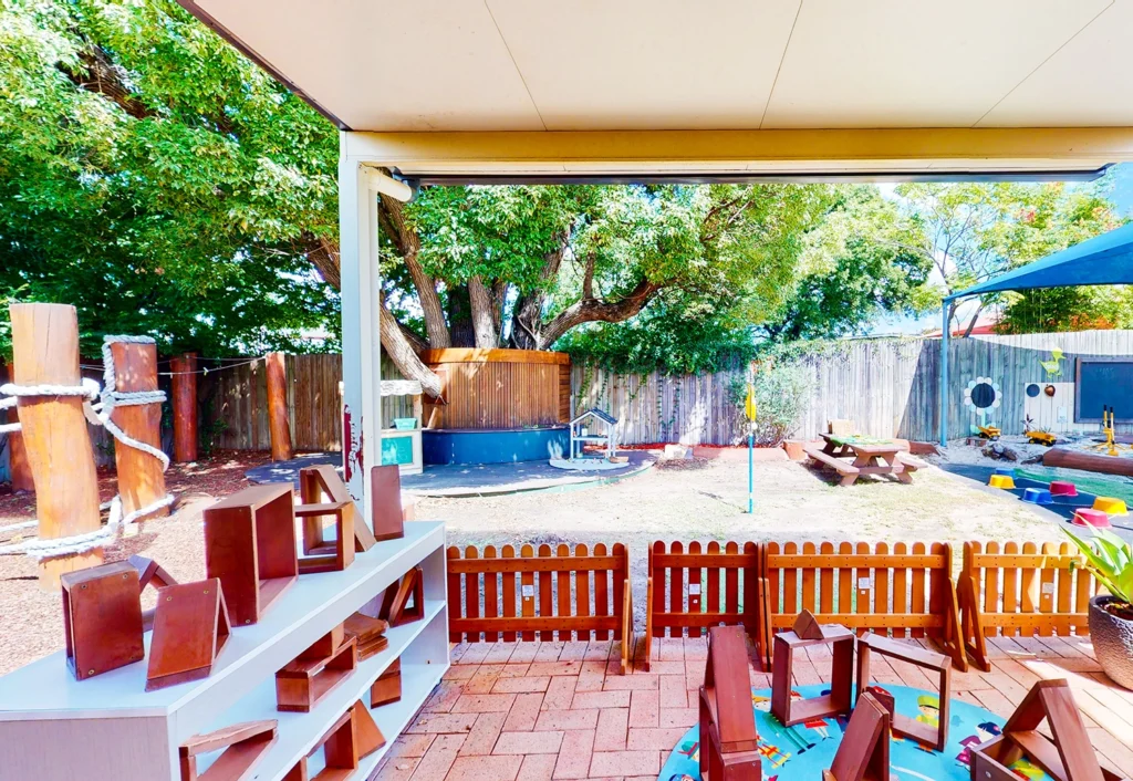 Outdoor daycare play area featuring wooden furniture, climbing equipment, and a shaded picnic table, surrounded by trees and a wooden fence. This engaging preschool environment is viewed from a covered patio area with brick flooring.