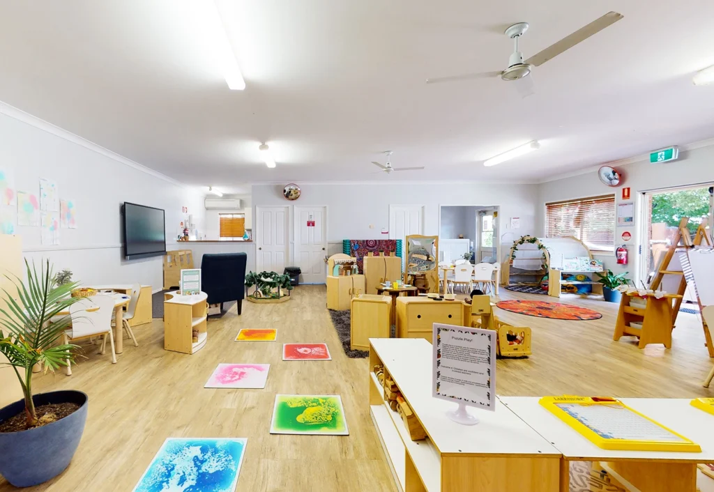 A brightly lit preschool classroom with various learning stations, children's artwork on the walls, educational toys, and furniture arranged neatly. Ceiling fans and indoor plants are also visible.