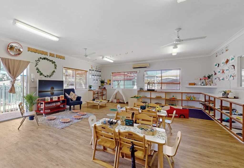 A spacious classroom designed for early learning, featuring wooden furniture, children's activity tables, chairs, a play area, a TV, and educational decorations on the walls. The room boasts large windows that allow ample natural light to fill this vibrant preschool environment.