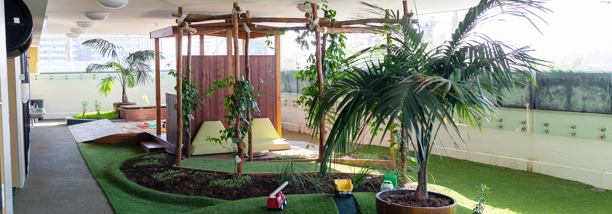 Indoor garden area with potted plants, a wooden pergola, lawn chairs, and children's toys on artificial grass. Perfect for early learning and preschool activities, walls are adorned with greenery while natural light filters through large windows.