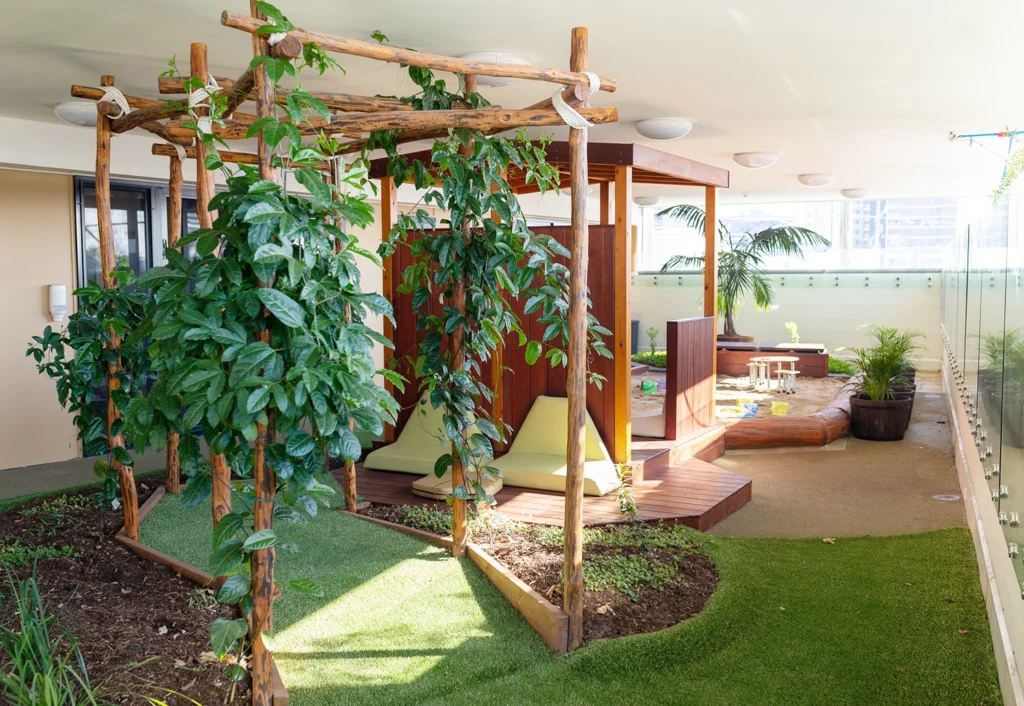 An indoor garden with lush plants, a wooden structure, seating area, sandbox, and glass walls allowing natural light creates a perfect early learning environment for preschool children.