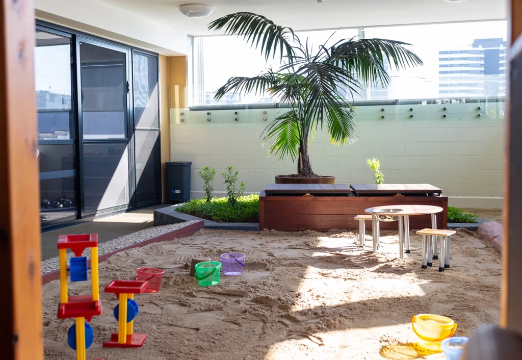 Indoor play area with sand, colorful toys, small tables and chairs, and a large potted palm ideal for preschool and early learning. Sunlight is streaming in through large windows.