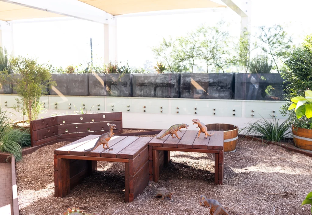 A landscaped outdoor play area for the daycare features wooden tables and benches, adorned with small dinosaur figurines and surrounded by lush greenery.