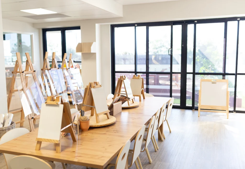 A well-lit art studio designed for early learning, with easels, drawing boards, and stools arranged around a large wooden table. Large windows in the background provide ample natural light for young artists in preschool or kindergarten.