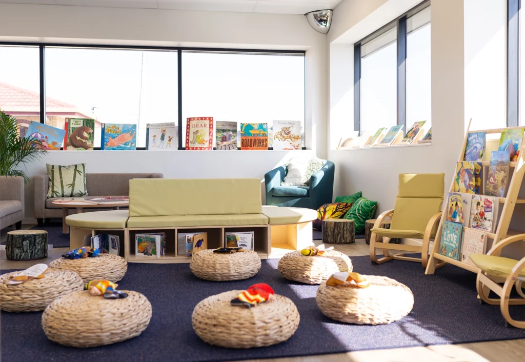 A brightly lit children's reading area at the preschool features rattan seating, colorful cushions, and shelves filled with books. Large windows allow natural light to flood the cozy space, creating an inviting atmosphere for early learning.