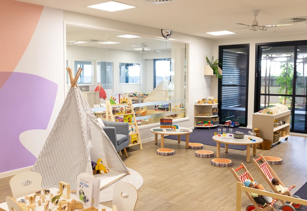 A brightly lit playroom designed for early learning with a teepee, various toys, bookshelves, and small tables. There's a large window and sliding doors, with wooden stools and colorful decorations perfect for kindergarten childcare activities.