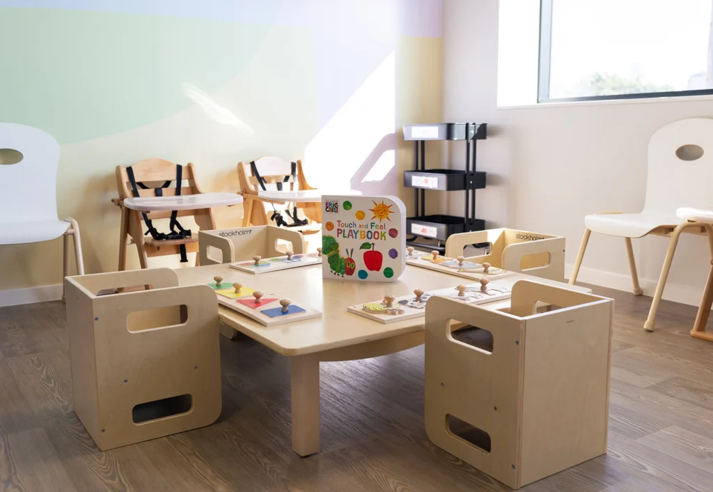 A brightly lit daycare playroom with a wooden table and chairs, featuring a "Touch and Feel Playbook" and various educational toys designed to enhance early learning.