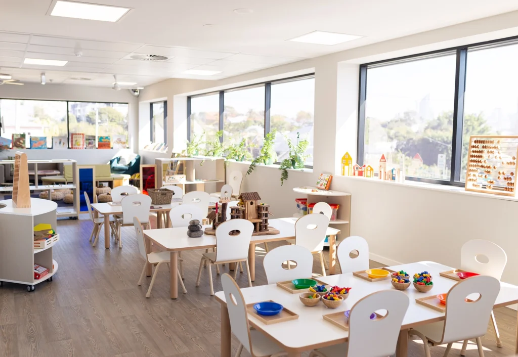 Bright and airy preschool classroom with white tables, small chairs, and shelves filled with educational toys and books. Large windows allow natural light to enter, creating a welcoming environment for children.