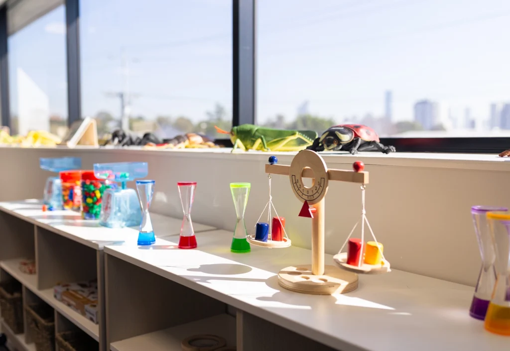 A sunlit classroom shelf displays colorful hourglasses, a wooden balance scale with weights, and various educational toys, creating an inviting space for kindergarten learning.