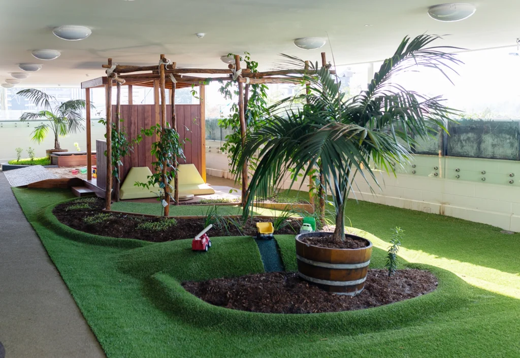 A modern indoor plant area with a wooden pergola, various plants, palm trees, and a wall-mounted purple screen perfect for early learning activities. Artificial turf covers the ground, creating an engaging space for little ones.