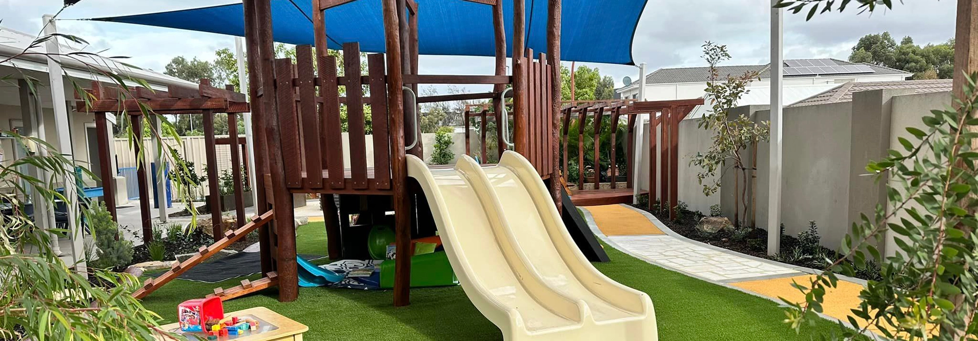 Outdoor playground with a wooden structure, two slides, a climbing area, and a shaded canopy. The surrounding area has artificial grass, plants, and a small table with toys.