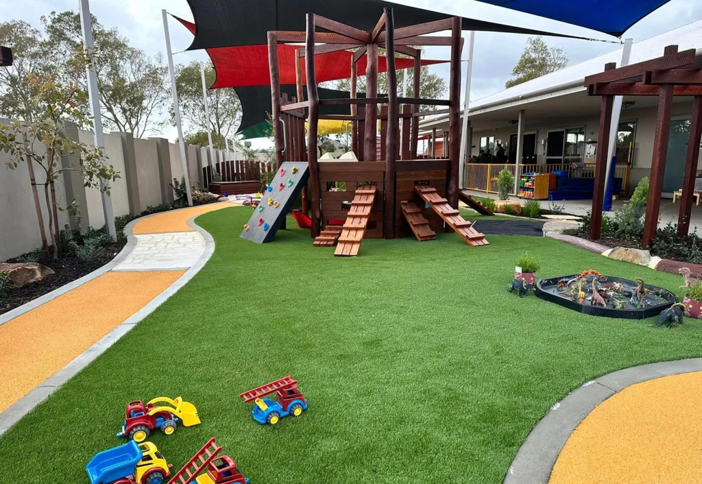 A colorful playground with artificial grass, a wooden climbing structure, slide, small toy vehicles, a pathway, and shade sails overhead. Trees and a white building are in the background.