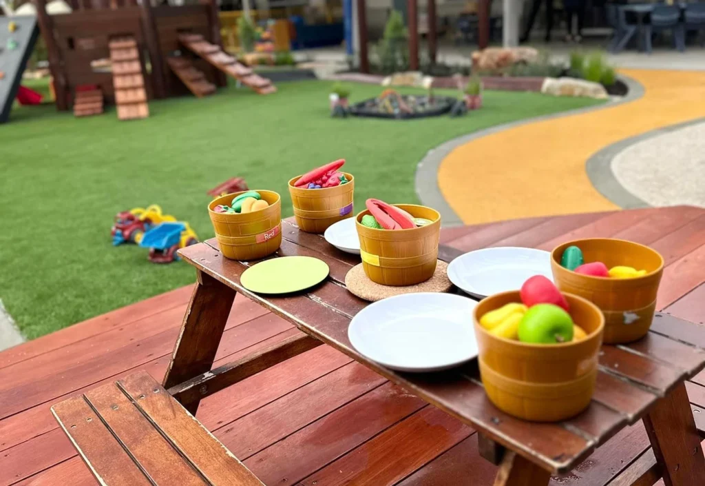 A picnic table with baskets of toy fruits and vegetables, toy plates, and a toy pot, set up in an outdoor children's play area with a playground in the background.