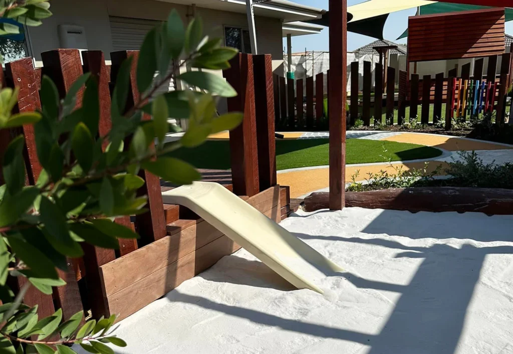 A small playground features a slide leading into a sandpit. Wooden fences and structures are seen, with a shaded area and artificial turf in the background. A leafy branch is in the foreground.