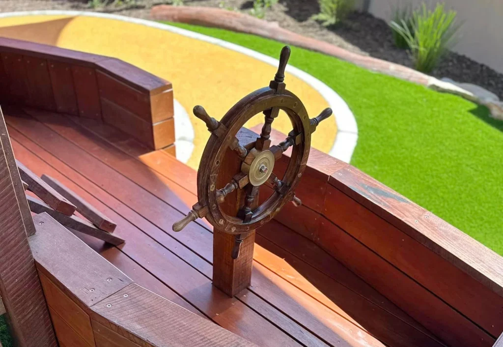 A wooden ship wheel is mounted on the deck of a wooden play structure, with artificial grass and a sandpit visible in the background.