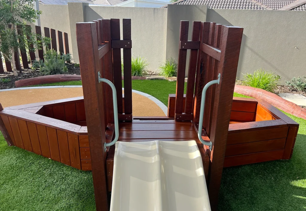 A small wooden playground structure with a white plastic slide extending downwards. The structure is surrounded by artificial green grass and the background shows a beige wall and some plants.