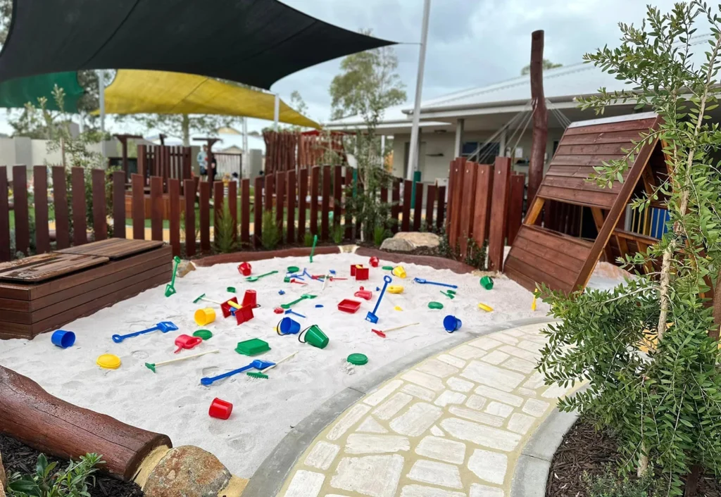 A sandbox filled with various colorful plastic toys is situated in an outdoor play area, surrounded by a wooden fence and greenery.