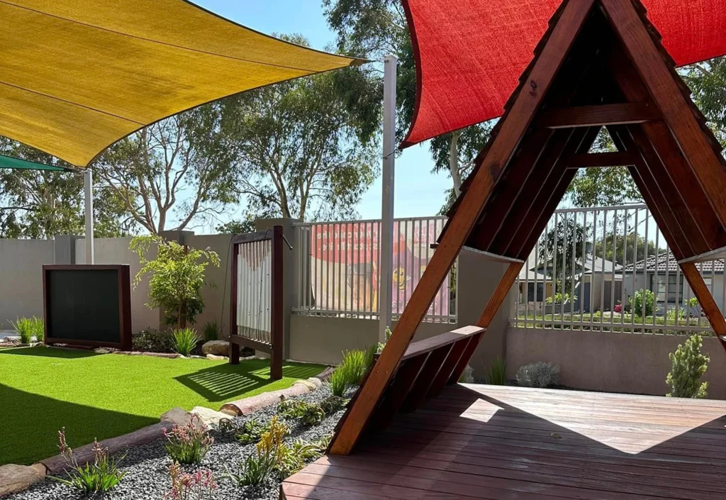 A backyard with two shade sails, one beige and one red. There is artificial grass, a small garden with rocks, and a wooden triangular structure on a deck. A railing and trees are in the background.