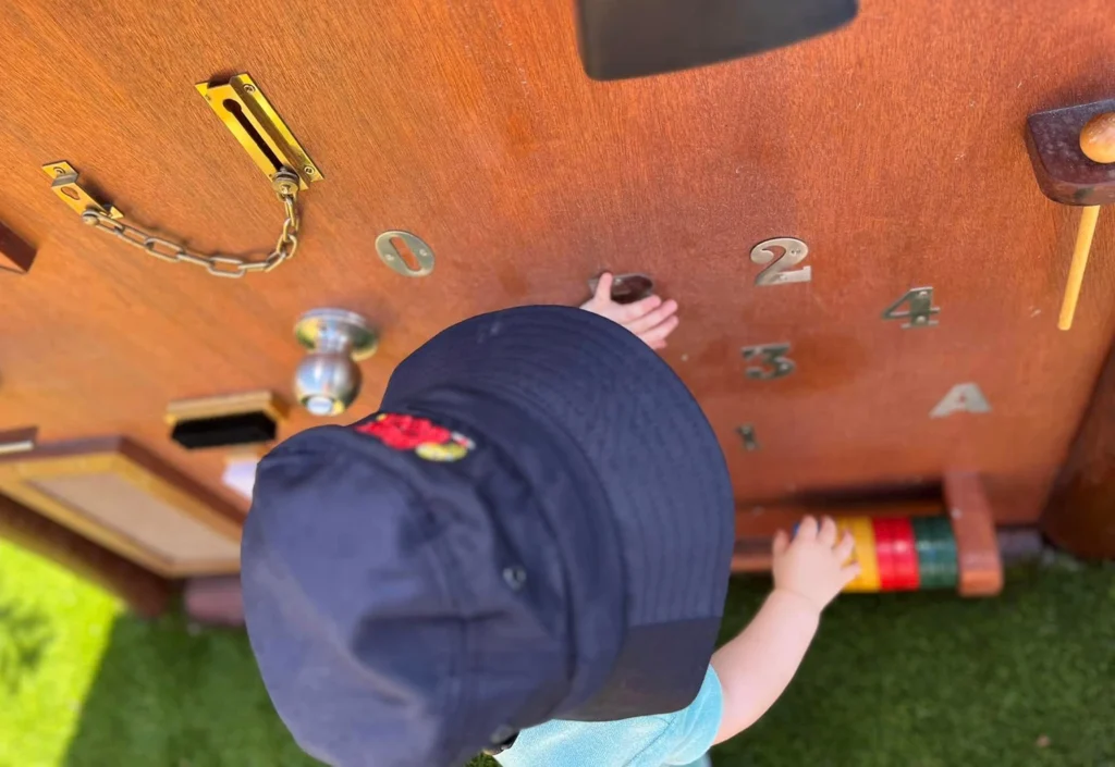 A child wearing a navy hat and light blue shirt plays with a wooden activity board featuring numbers and various latches outside.