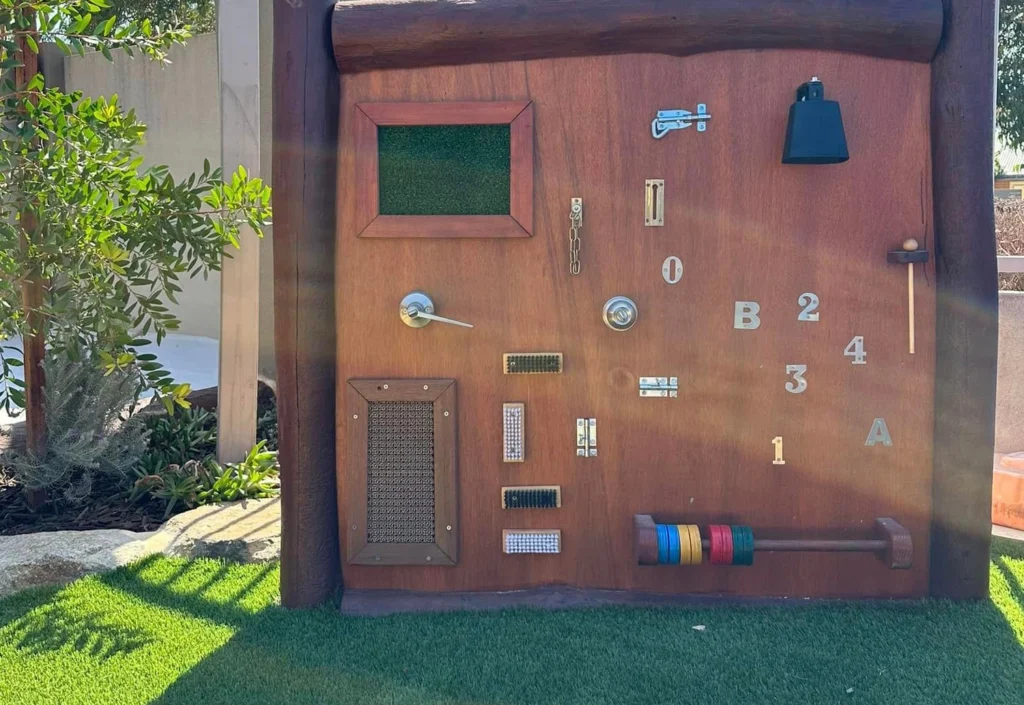 A wooden activity board stands in a garden with various latches, locks, handles, and gears for children to interact with, against a backdrop of greenery and sunlight.