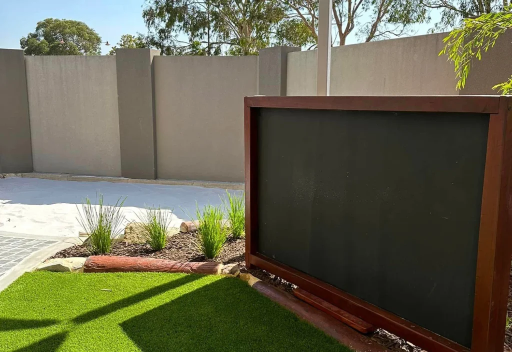 A backyard with artificial grass in the foreground, a planted flower bed, a large empty blackboard, and a high concrete fence in the background on a sunny day.