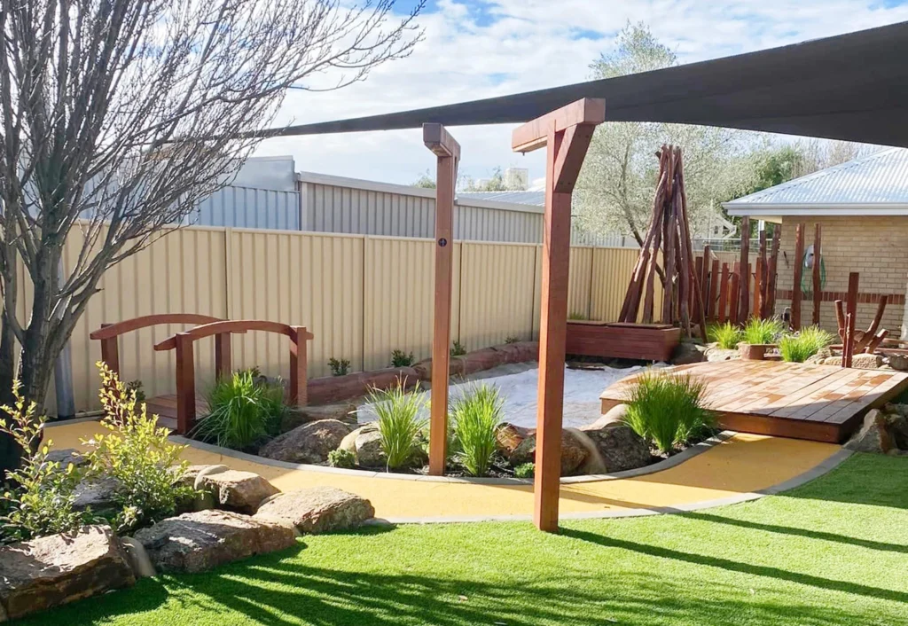 A landscaped backyard with a winding pathway, wooden bridge, shaded seating area, and green lawn surrounded by plants and rocks. Fence lines the edge, and trees are visible in the background.