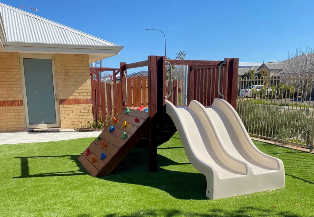 A small playground with dual slides, a rock climbing wall, and artificial grass surface, located next to a building and fenced area.