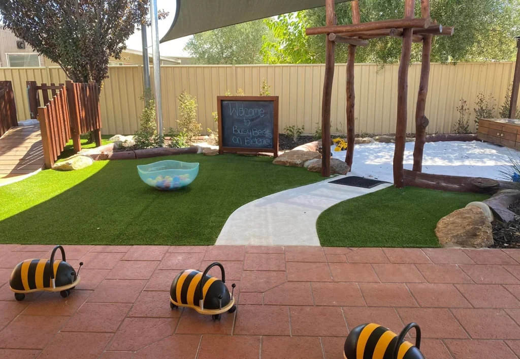 A children's outdoor play area with artificial grass, a paved walkway, a chalkboard with "Welcome," and bee-shaped toys on the ground. A sandpit and wooden climbing structure are in the background.