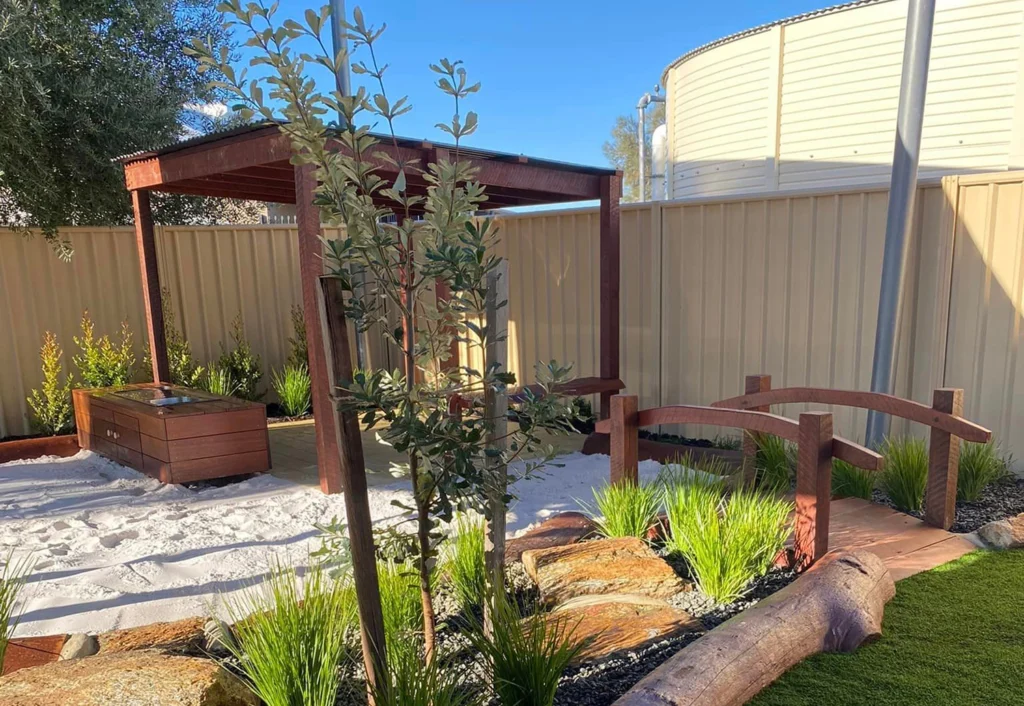 A small garden with a wooden pergola, a sandbox, a wooden bench, a small arched bridge, and various plants, framed by a beige fence and a large water tank in the background.