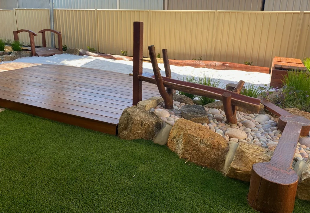 A backyard features a wooden deck, artificial grass, a white sand area, a small wooden bridge, and decorative rocks integrated with wooden log seating and landscape elements.