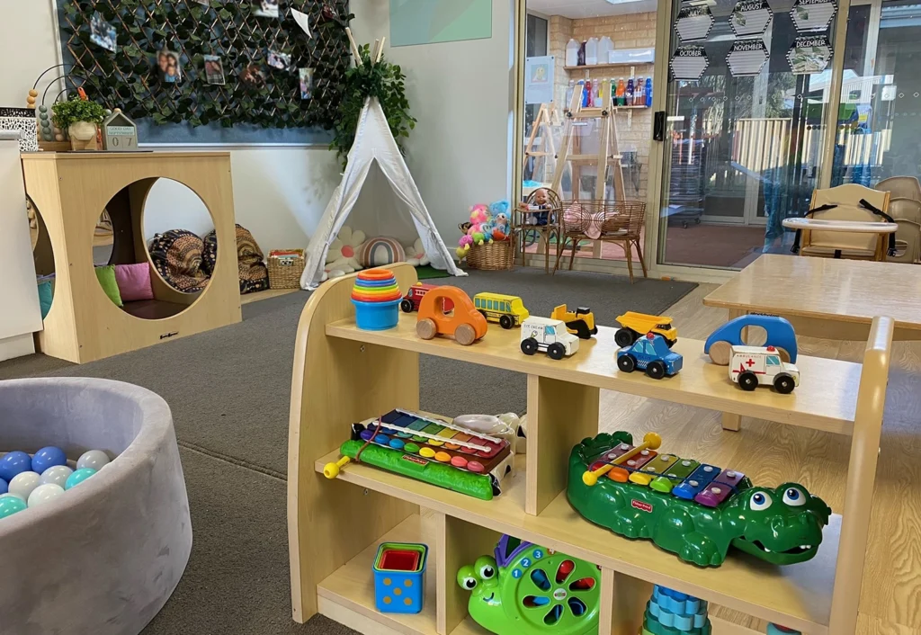 A brightly lit playroom with shelves of toys, a ball pit, a teepee, and various activity centers. There are tables, chairs, and colorful decorations throughout the room.