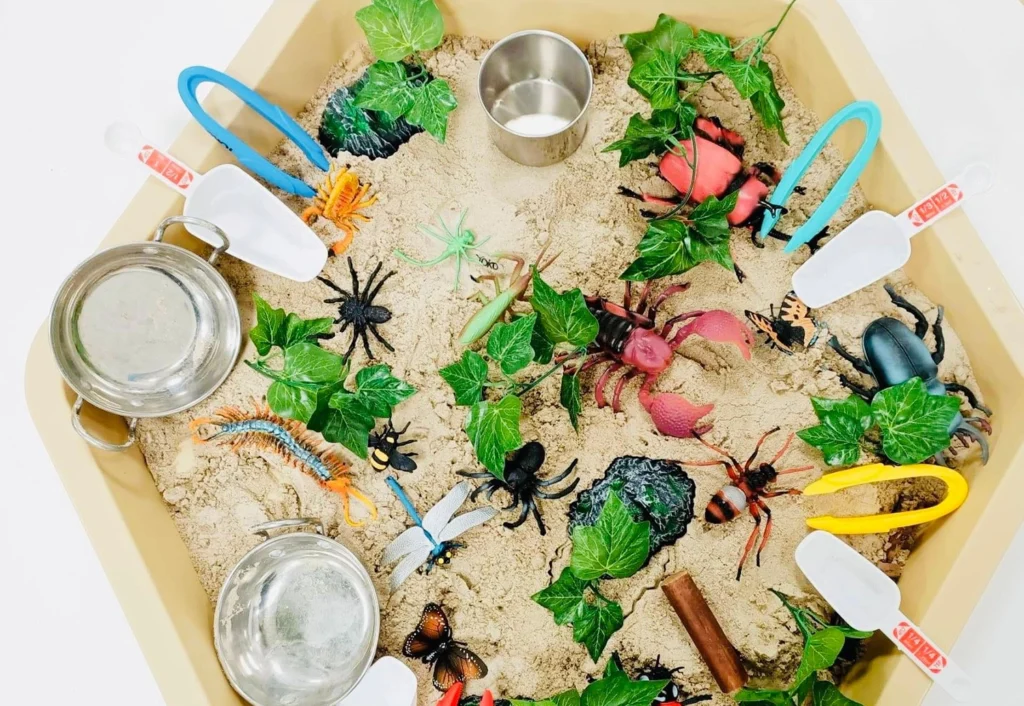 A sensory play tray filled with plastic insects, leaves, rocks, and sand. Various tools, including scoops and tongs, are placed around the tray.