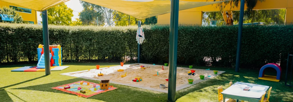 Outdoor children's play area featuring a large sandpit with various toys, a colorful play structure, a small table with chairs, and a play tunnel. Shade cloths are seen overhead, making it an ideal spot for early learning and playful adventures.