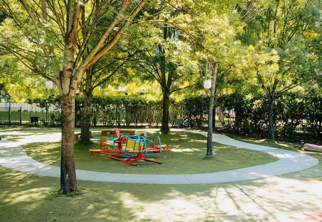 A small playground at the day care features colorful equipment surrounded by trees and shaded by green foliage. A curved pathway encircles the play area, offering a perfect spot for early learning adventures.