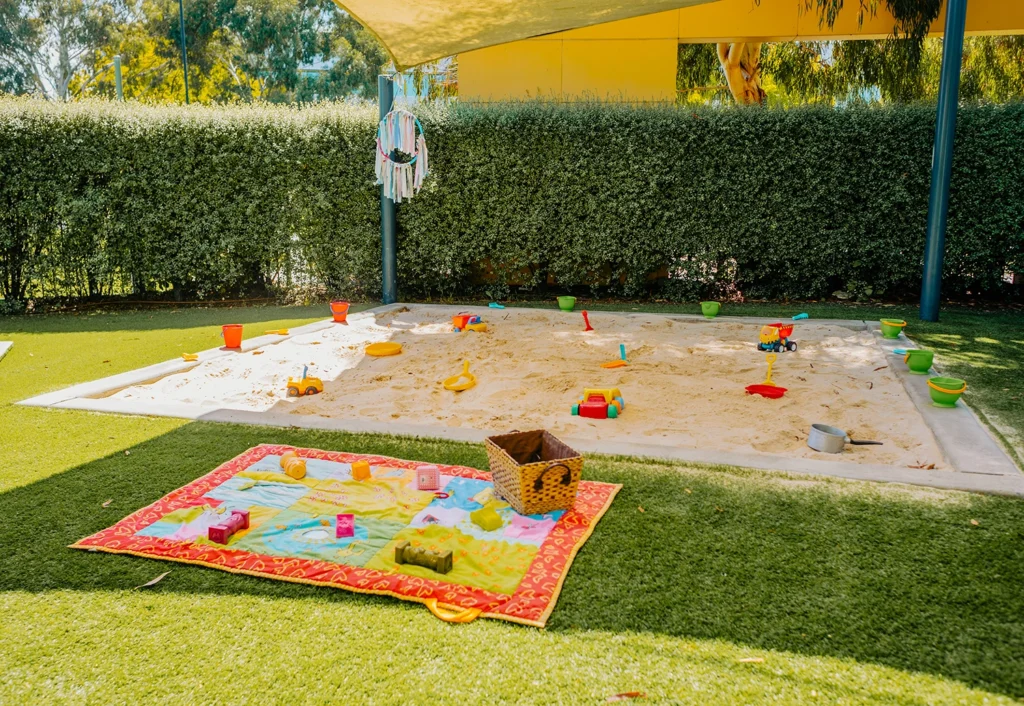 A sandy play area with toy shovels, buckets, and a blanket with toys laid out on the grass nearby. A hedge and a shade structure surround the play area—a perfect spot for any preschool or day care activities.