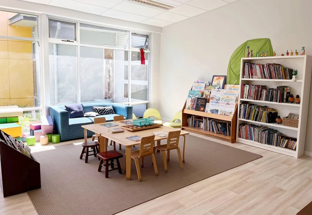 A brightly lit children's reading area in the preschool features a table with small chairs, shelves filled with books, a blue sofa, and colorful seating cubes near a window.