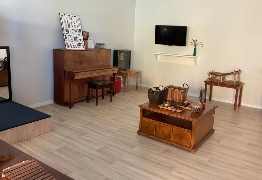 A clean, bright room featuring a wooden piano, coffee table with wicker basket, wall-mounted TV, and various decorative items—ideal for an early learning environment.